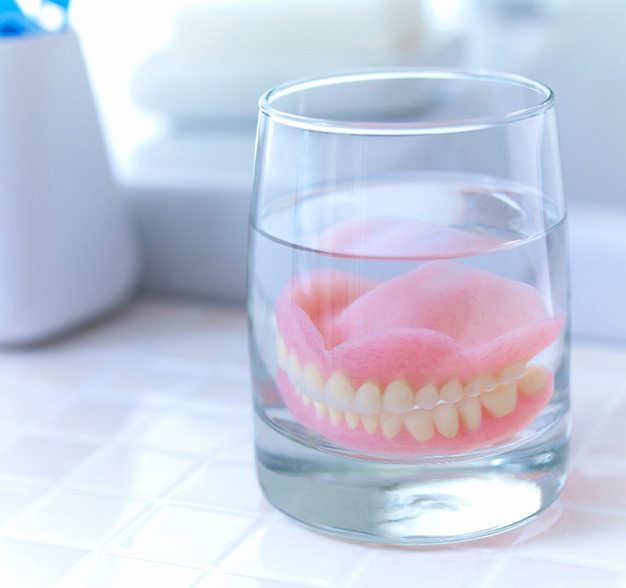 Complete set of dentures in clear glass of liquid on tiled surface