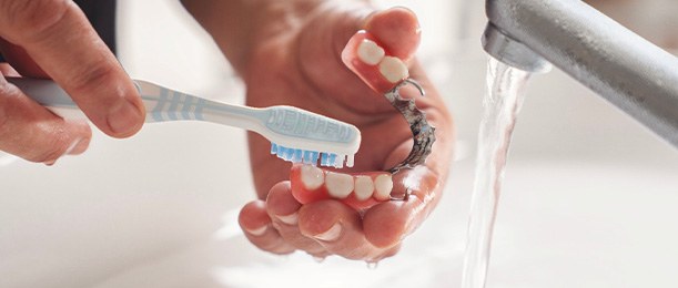 Hands brushing partial dentures at sink with running water