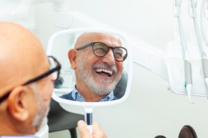 Older man with dental implants sitting in dentist’s chair smiling while looking into handheld mirror.